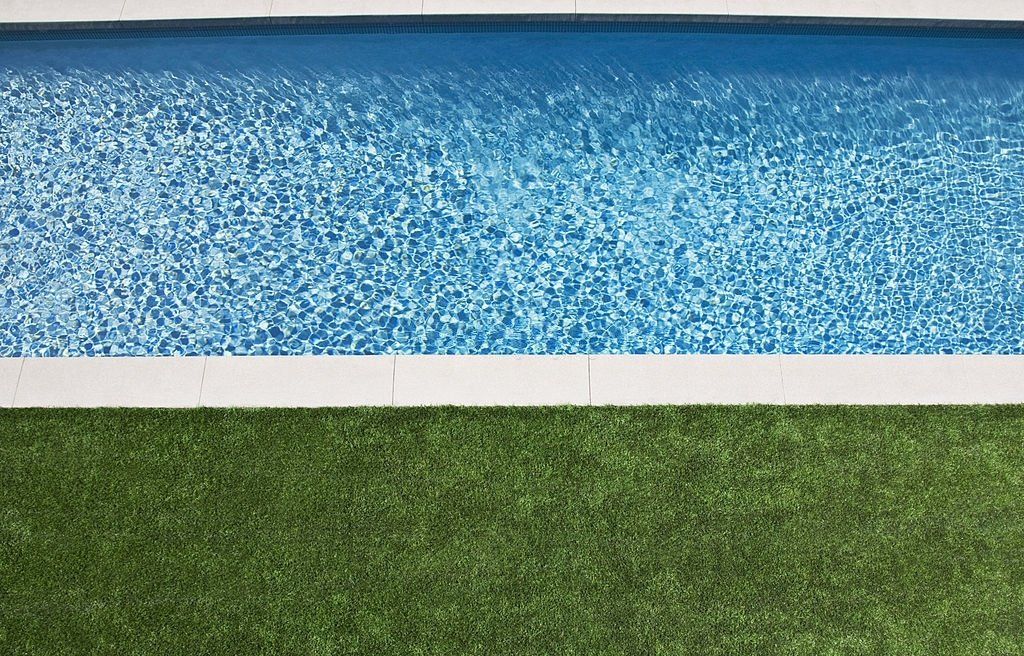 A serene swimming pool with clear, sparkling blue water is bordered by white tiles. Adjacent to the pool, there's a lush green grass lawn from one of the top Artificial Grass Installers in Jacksonville, FL, creating a vivid contrast between the cool blue water and the vibrant greenery.
