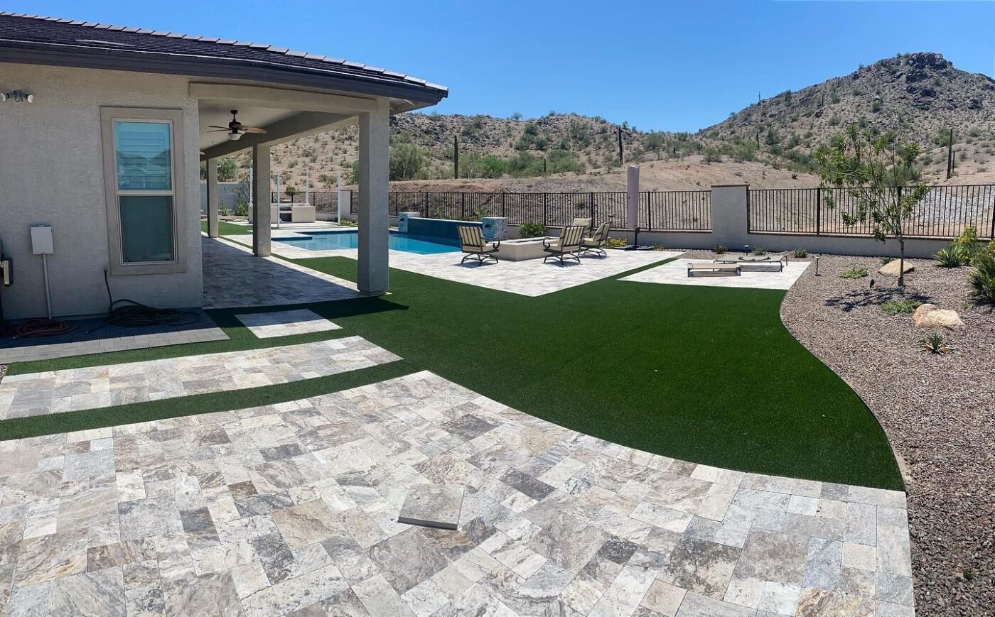 A spacious backyard featuring a large tiled patio and an artificial turf area. This inviting outdoor space includes a covered patio with a ceiling fan on the left, leading to a pool with a sun deck and lounge chairs. The background shows a fenced area with desert landscaping and a mountain view.
