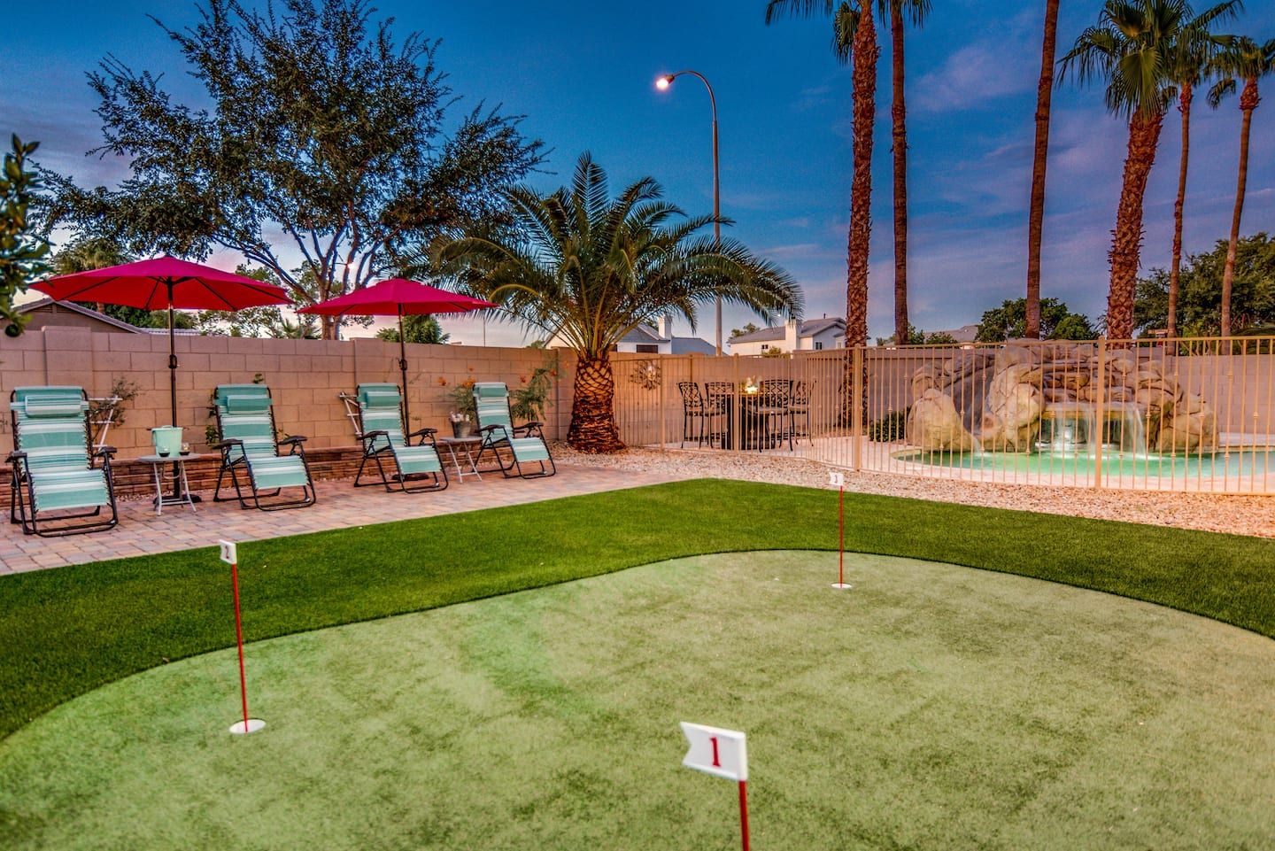 A backyard scene at sunset featuring a small putting green with numbered holes, meticulously laid by top artificial grass installers. Several lounge chairs with side tables and red umbrellas are near a wall adorned with palm trees. In the background, a lit pool area with rocks and additional palm trees completes this Jacksonville, FL oasis.