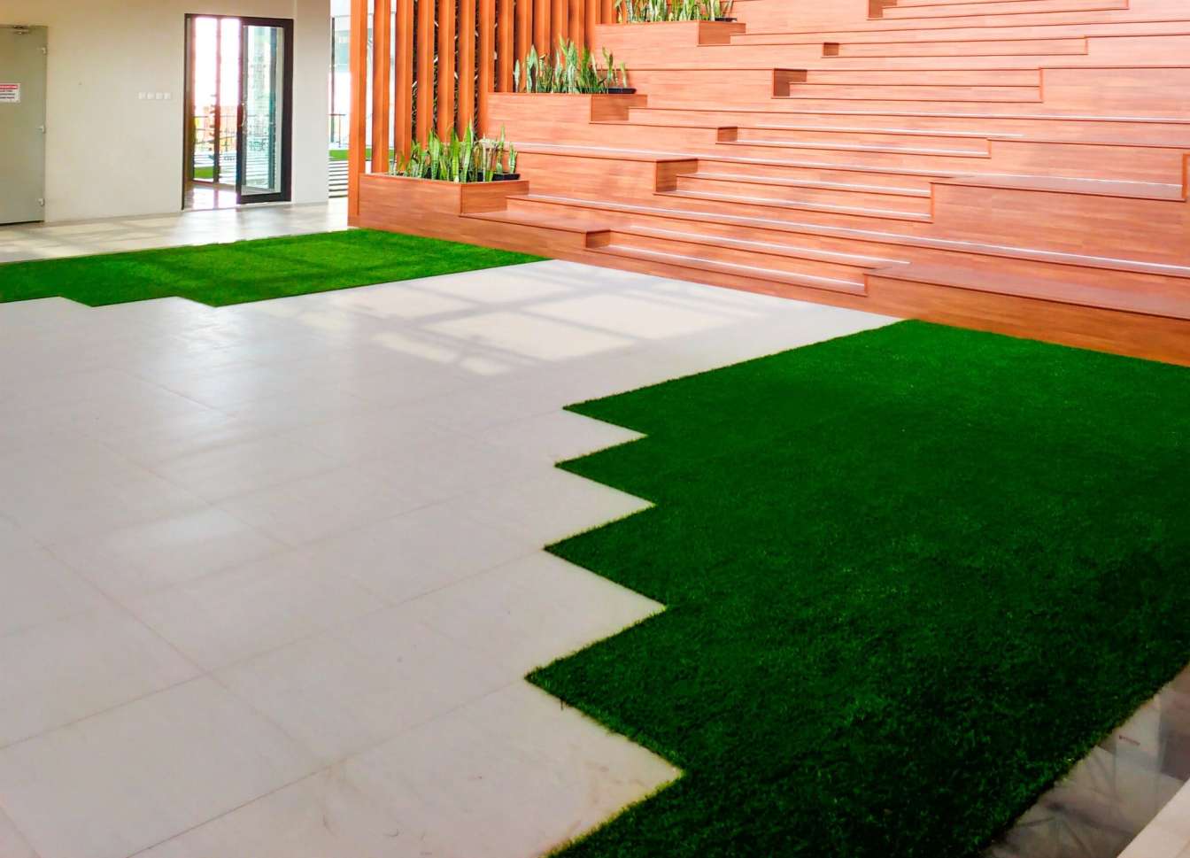 A modern interior space in Jacksonville, FL, featuring wood panel steps with integrated planters on the right. Adjacent to a pattern of green artificial grass and white tiles on the floor, this area is well-lit with large windows visible in the background—perfect for those seeking expert turf installations.