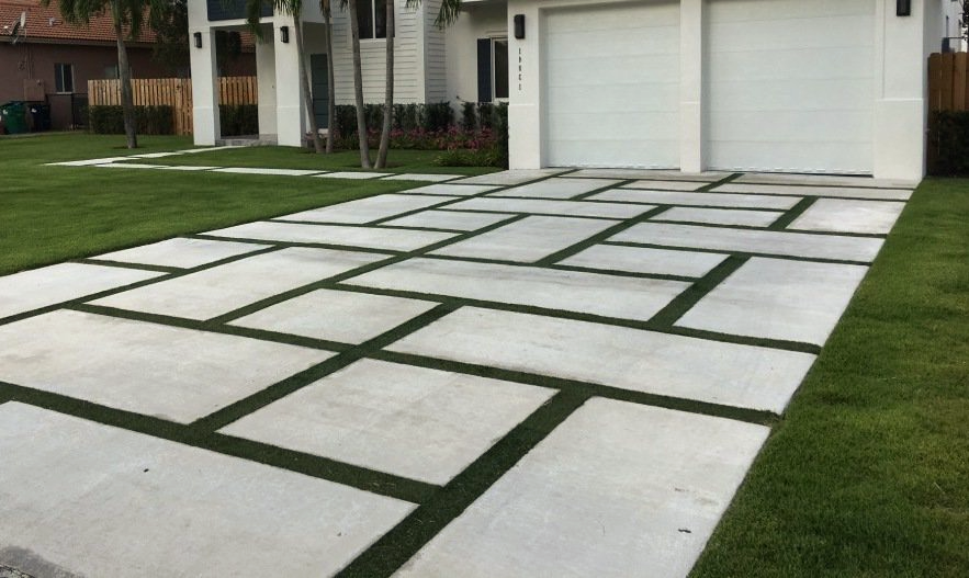 A modern driveway with a unique geometric design features large rectangular concrete slabs interspersed with strips of lush artificial grass. Ideal for turf installations in Jacksonville FL, the driveway leads up to a double garage with white doors, and the adjacent lawn is well-maintained.