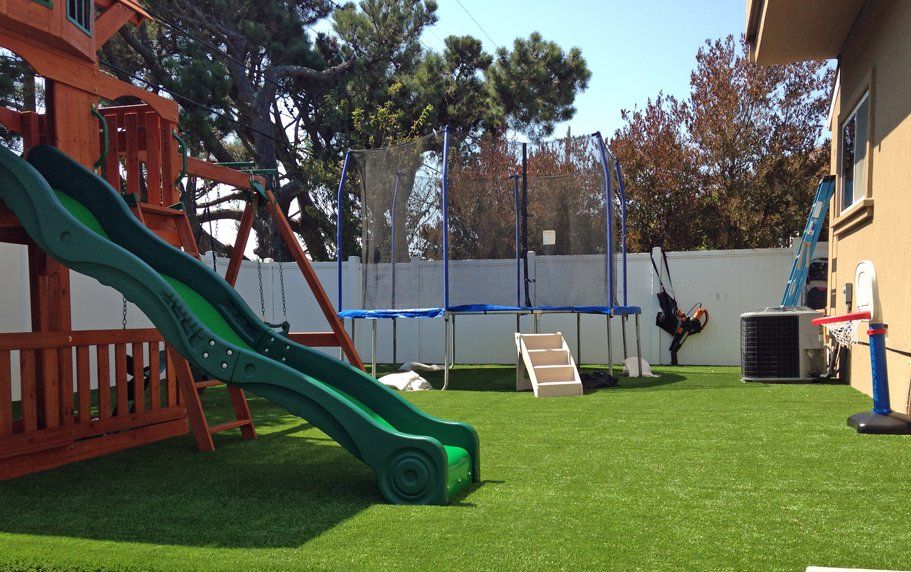 A backyard with a green slide attached to a wooden playset, a trampoline with a safety net, a swing, and a basketball hoop. The area features artificial grass installed by professional turf installations in Jacksonville FL and is enclosed by a white fence. Trees are visible in the background.