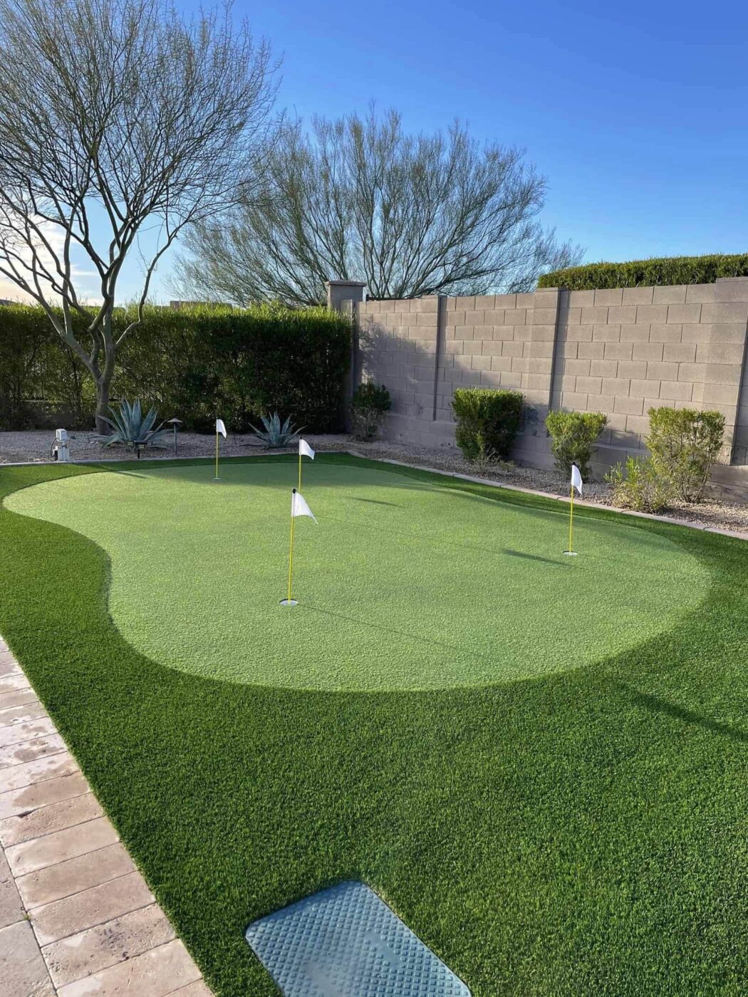 A backyard scene at sunset featuring a small putting green with numbered holes, meticulously laid by top artificial grass installers. Several lounge chairs with side tables and red umbrellas are near a wall adorned with palm trees. In the background, a lit pool area with rocks and additional palm trees completes this Jacksonville, FL oasis.